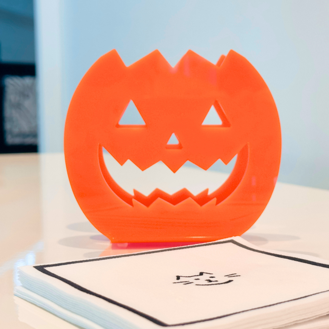 Orange jack-o-lantern napkin holder with a stack of white napkins with a small black cat design on a white background 