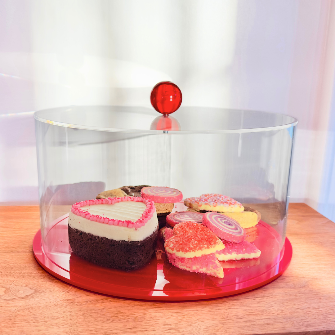 Clear cake stand with red base holding heart-shaped cookies on a wooden surface.