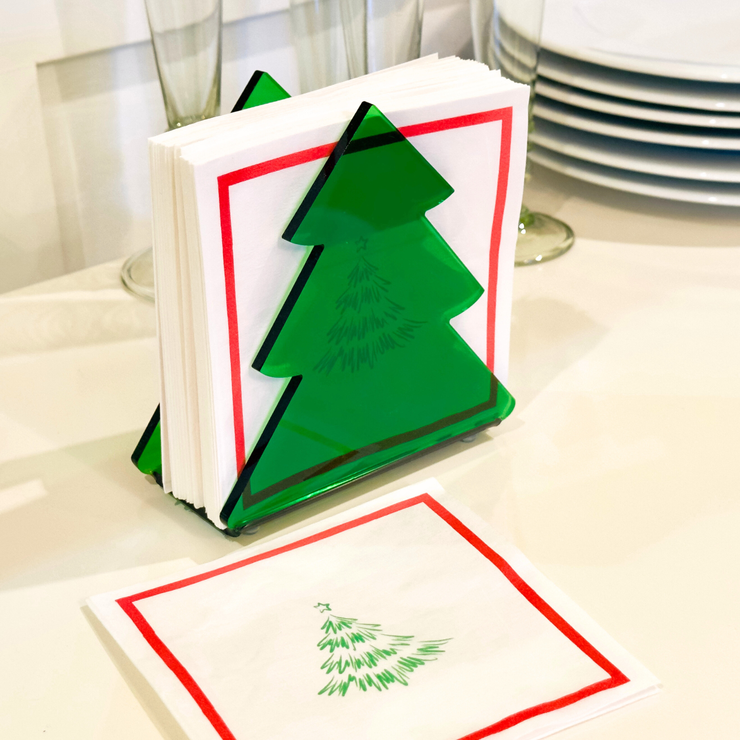 Stack of white napkins with a green tree illustration on top inside a green Christmas tree holder on a table.