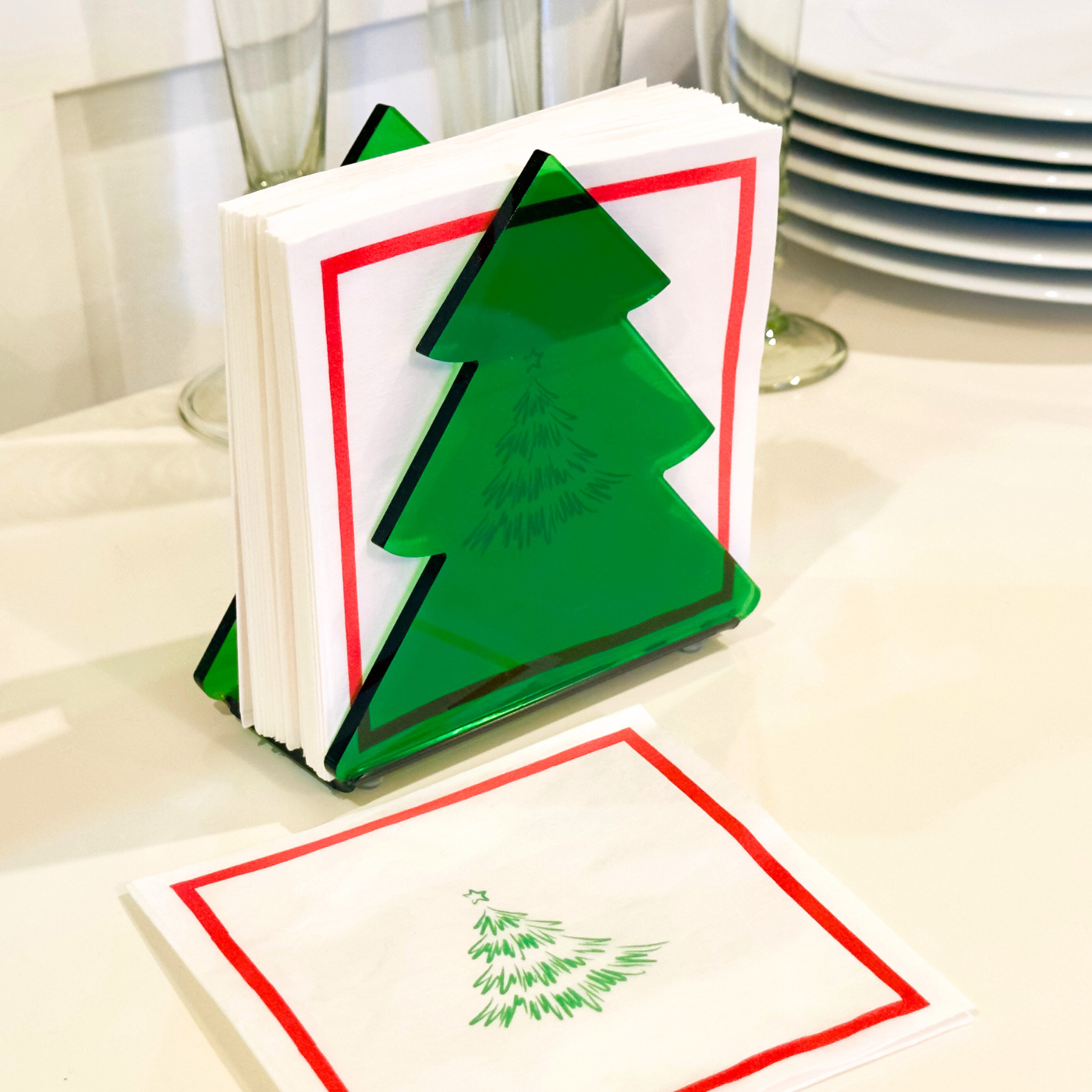 Stack of white napkins with a green tree illustration on top inside a green Christmas tree holder on a table.