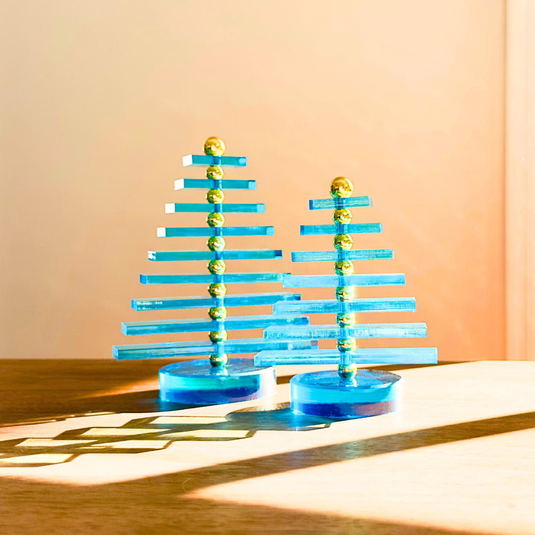 Two blue Christmas tree figurines with gold balls on a wooden surface and peach-colored wall background.