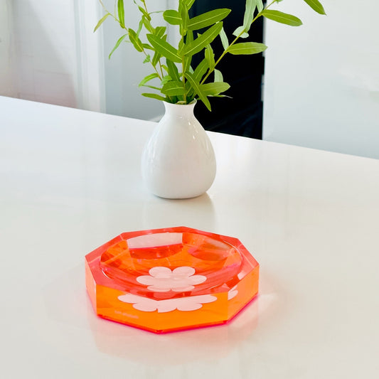 Hexagonal orange dish with floral design on a white surface, next to a white vase with green leaves.
