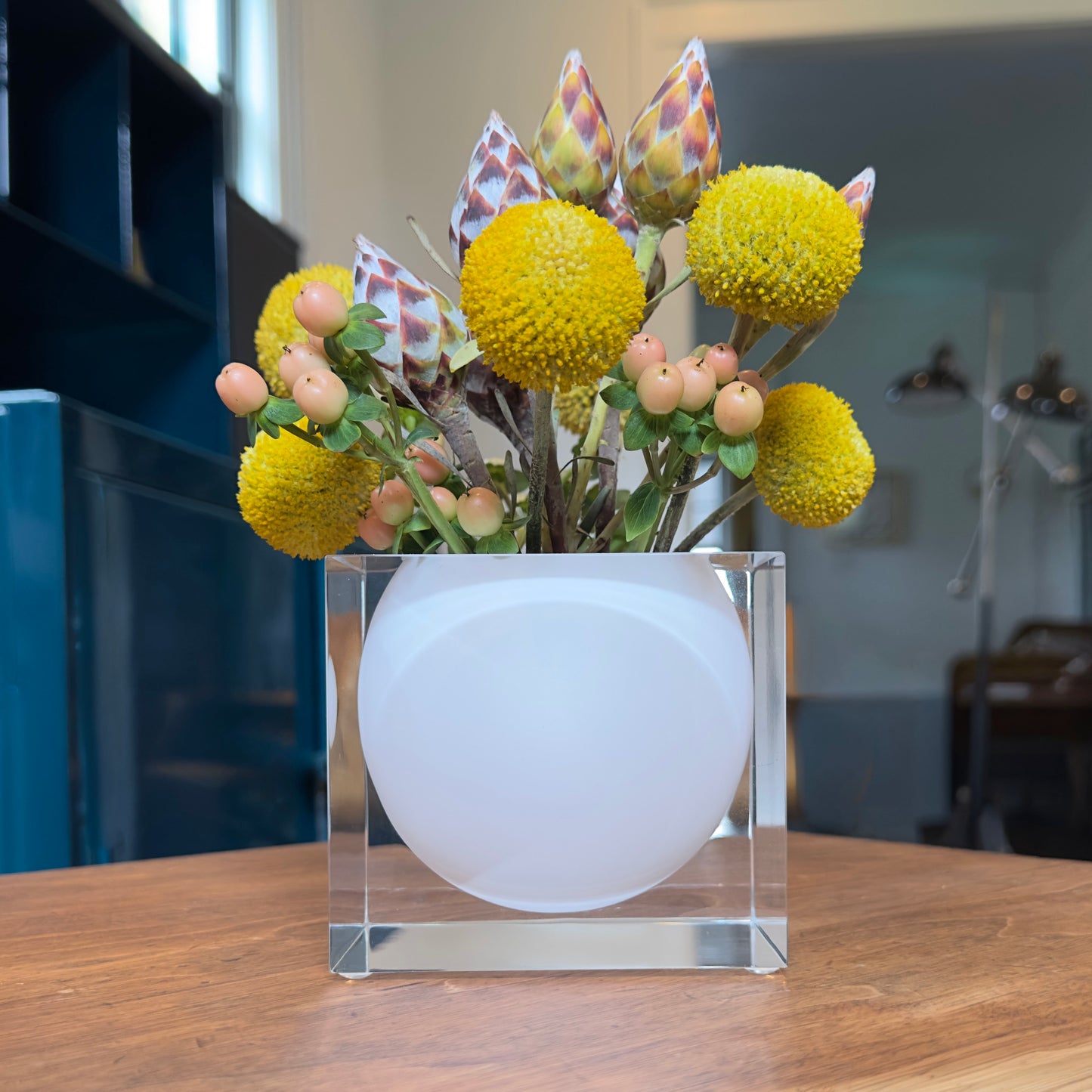 Clear cube with white vase inside with yellow flowers on a wooden surface