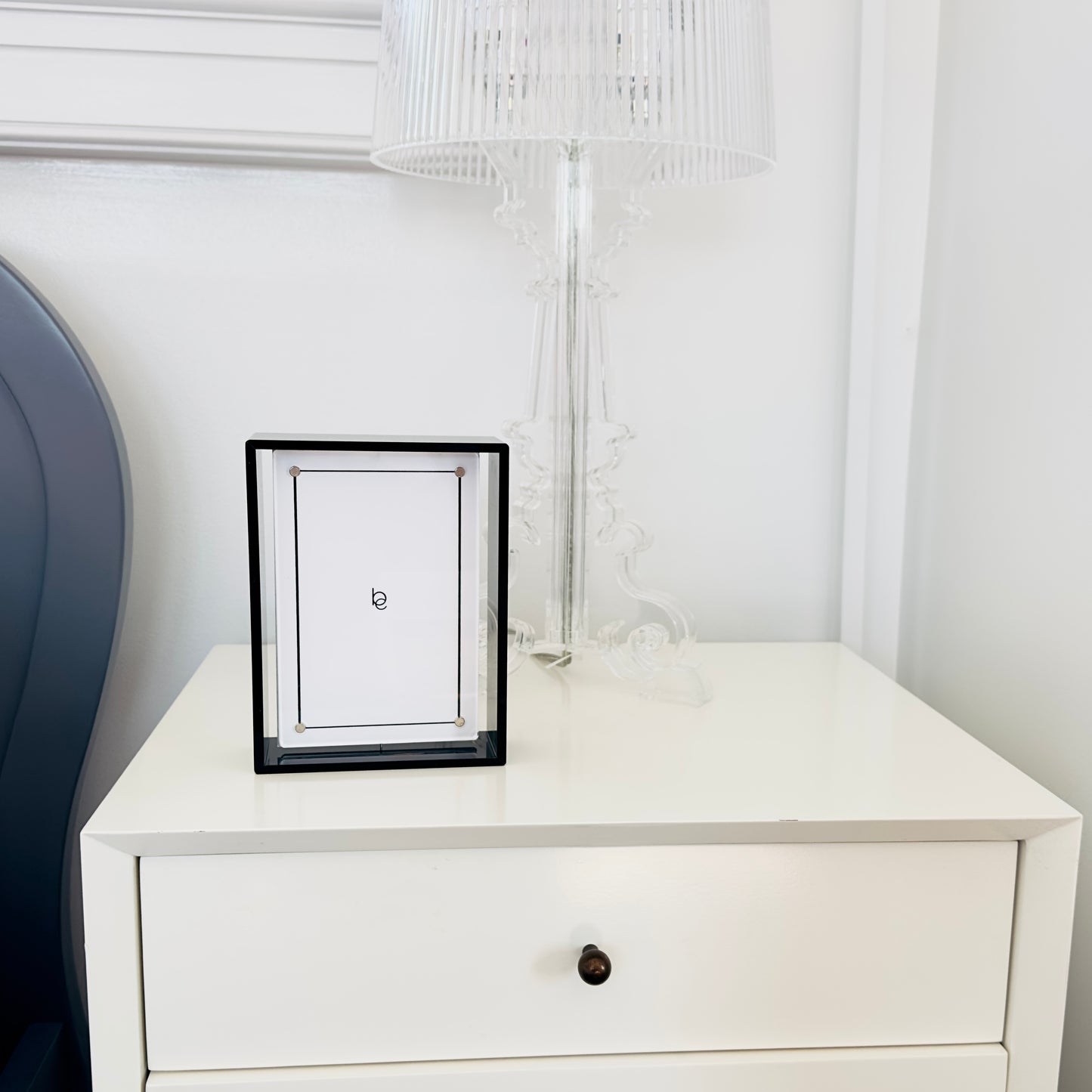 White nightstand with a framed picture and lamp against a white wall.