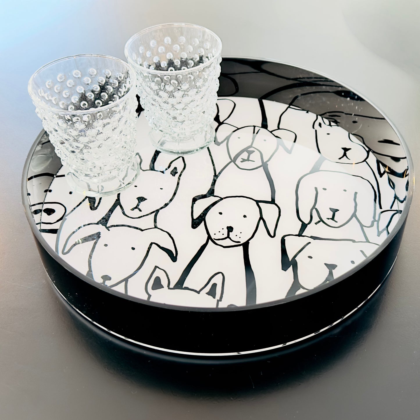 Round tray with black rim featuring black and white dog illustrations, topped with two small glass cups.