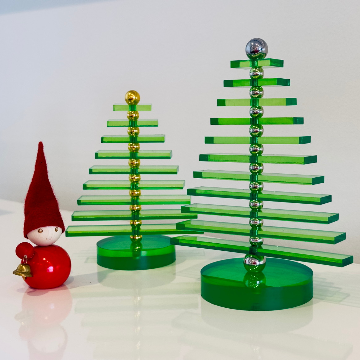 Two green acrylic Christmas trees with gold and silver accents next to a red gnome figurine on a white background.