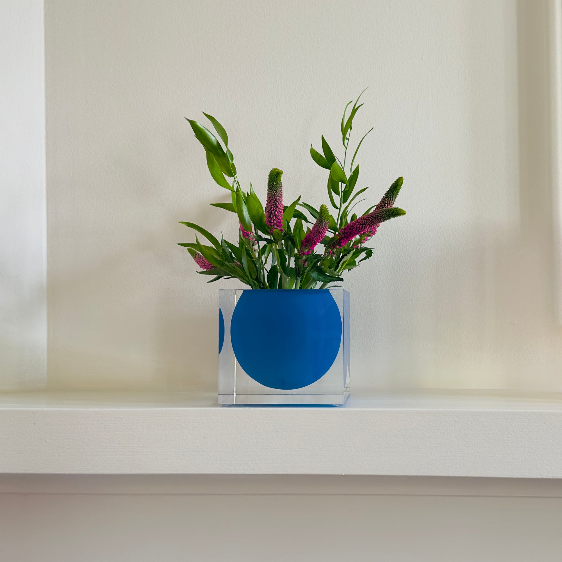 Blue acrylic vase with green leaves and pink flowers on a white surface