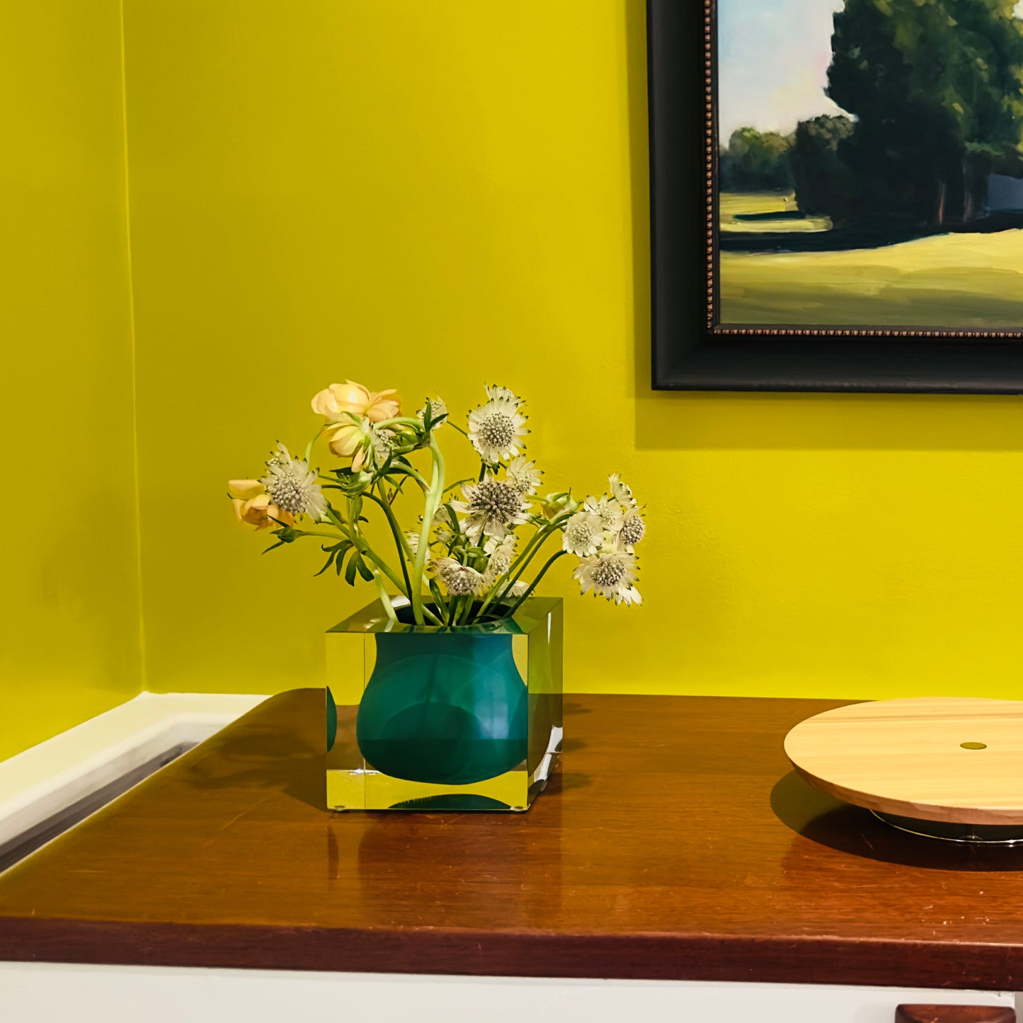 Green vase with flowers on a wooden surface against a yellow wall with a framed picture.